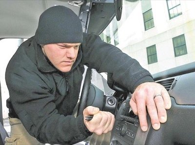 A person wearing dark clothing reaches through a broken car window, attempting to steal a car stereo system. The image illustrates vehicle break-in and theft.
